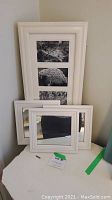 Large rectangular white picture frame with four black and white floral prints leaning against wall beside two smaller square white framed mirrors.