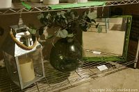 Photo showing metal lantern with thick rope handle containing fake candle, glass vase with artificial greenery, and mirror tray on a wire shelving unit.