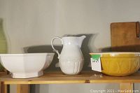 Wide shot showing large white square bowl, white pitcher with embossed detail, and part of the yellow round bowl on a wooden shelf