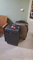 Two large hard shell suitcases, one black and one bronze/tan color, positioned on floor next to a chair and carpet.