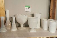 Front view of assorted milk glass items including large scalloped pedestal bowl, smaller pedestal bowl, various vases including tall and ribbed designs, and five hobnail cups.