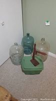 Three large glass wine jugs and a red manual corker on a green plastic base, positioned in corner of room.