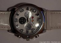 Close-up photo of a quartz wristwatch with a white leather band and silver-tone round case, showing the dial with Arabic numerals, three subdials, and decorative dots.