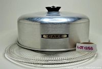 Aluminum cake carrier with domed lid, black knob, and 'CAKE' label on front, accompanied by round patterned glass plate beneath.