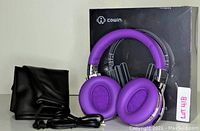 Photo shows new purple Cowin E7 headphones with cushioned ear pads and headband, black carrying pouch beside them and original black box packaging in background.