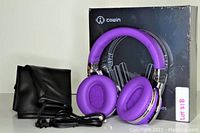 Purple Cowin E7 over-ear headphones displayed with black carrying pouch, audio cable, and the product box in the background.