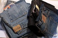 Photo shows folded jeans in a pile with visible back pockets, brand leather patches, and tags. Different denim colors visible including light blue, dark blue, and black.