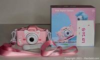 Pink kids digital camera with white lens, pink adjustable strap, and retail box showing pink and blue camera images.