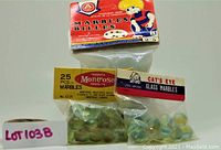 Three sealed packs of glass marbles, labeled as Montrose 25 pieces, Cat's Eye glass marbles, and an additional pack with a red box labeled 'Marbles Billes' featuring a cartoon character.