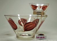 Two glass cups, one larger and one smaller, both hand-painted with brown leaf designs. The smaller cup is held in a gold-tone metal holder with a handle.