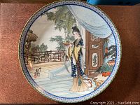 Top-down photo clearly showing the whole plate with intricate traditional Chinese painting of a woman standing in a veranda with plants and architectural details, signed with calligraphy, plate rim decorated.
