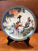 Front of the porcelain collector plate with colorful detailed traditional Chinese painting of a woman sitting at a table among plants