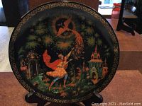 Photo of black lacquered round decorative plate featuring vibrant traditional painting of the Tsarevich reaching for the Firebird amid fantasy foliage and buildings.