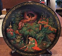 Full view of the round collector plate on a stand showing the detailed Firebird story scene painting and gold ornate border on dark background.