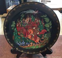 Front of the porcelain collector's plate with vibrant, detailed painting depicting a knight on horseback and a red castle surrounded by trees, framed with a gold ornamental border on black background.