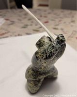 View of sculpture from the side showing the figure holding a long stick or rod in a kneeling position, highlighting the mottled green and black stone texture.