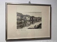 Framed black and white sketch artwork showing buildings along a river with people figures on the riverbank, signature at bottom right and title at bottom left.