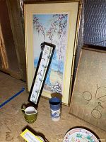 Four framed Asian style art paintings leaning against a wall with a small ceramic cup and creamer, and a tall blue and white ceramic container in front.