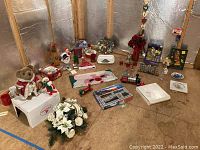 Photo shows a variety of Christmas-themed collectibles, figurines, floral arrangements, a teddy bear, tea sets, platters, a book, train set in box, and decorative items arranged on a plywood floor against foil insulated wall.