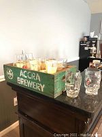 Overview of green wooden crate filled with assorted vintage beer and bar glasses, showing crate label 'Accra Brewery'.