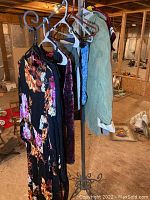 Overview of five women's party dresses, jackets and shirt hanging on a wrought iron clothes rack in an unfinished basement