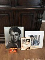 Wide shot showing the four memorabilia items: one large framed black and white Elvis photo, one smaller framed photo, hardcover book, and boxed card set in front.