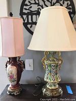 Both lamps shown side by side on a dark surface beneath a gray wall with decorative wall art. Left lamp has maroon urn shaped base with floral painted front, pink fabric rectangular shade. Right lamp has intricate multicolor enamel floral openwork ceramic base with white shade.