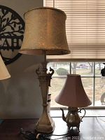 Tall vintage lamp with beige paisley fabric shade, and smaller silver-plated teapot lamp with pink shade side by side on a wooden table in front of window.
