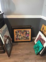 Seven framed art pieces and a wooden lobster pot wall hanging arranged on floor against wall, showing variety of sizes and colors.