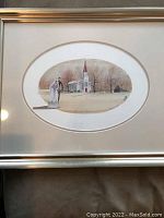 Photo showing entire framed print of bride and groom outside a white church with red spire, with oval mat and gold frame.