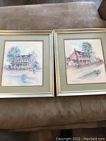 Two vintage framed paintings side by side on a couch, showing the full view of both artworks framed in silver-tone frames with green matting.