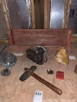 Wooden barn wood shelf, tin candle holder, slate and wood hatchet, temple bell, rustic metal cake mold, horseshoe paperweight, rustic vase all arranged on plywood surface