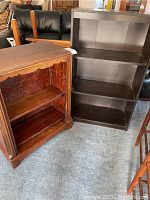Two bookcases side by side on floor. Taller bookcase painted dark brown with three shelves and no visible backing. Smaller hardwood bookcase with natural finish and two shelves, visible scratches and chips.