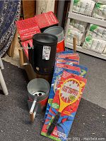 Photo showing three silver metal charcoal chimney starters and piles of packaged yellow bug zappers with wooden handles visible.