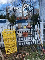 Outdoor black metal wire shelf unit with 5 shelves holding garden pots and other items, yellow plastic crates stacked to the left, garden tools leaning on the right, white picket fence background