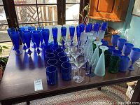 Photo of a large variety of colorful glassware on a table including blue tumblers, green tumblers, clear martini glasses, frosted glass Pilsner glasses, and stemmed glasses in blue and frosted colors.