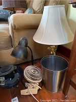 Two table lamps, a white clip-on fan, and a silver wastepaper basket grouped together in a room corner near a beige upholstered chair.