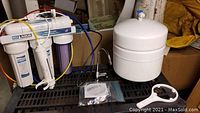 Full reverse osmosis system with three filtration housings attached to frame, plus pressurized white storage tank and assorted connectors packaged, all placed on black surface.