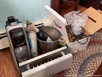 Sodastream machine, two gas canisters, silver ice bucket, black metal wine rack, and toaster boxed and arranged on wooden floor against wall.