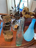 Photo showing clear textured glass vase, matte blue vase, vintage metal sculptures, teddy bear on wooden school desk, and metal container on a wooden table.