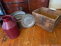 Four vintage items on wood floor: wooden box, metal pail, enameled jug, and metal plate.