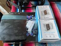 Black Cisco Linksys router and two boxed RangeXTD extenders with a white power plug extender visible on a colorful striped cloth surface.