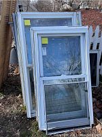 Five stacked white vinyl double hung windows leaning outside against a house and fence, some with screens visible. Outdoor storage with dirt and wear visible.