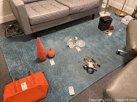 Blue textured area rug laid on floor with various small items placed on top for size reference, including orange bag and two decorative vases.