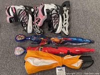 Photo showing two pairs of children's rollerblades and three umbrellas arranged diagonally on carpeted floor.