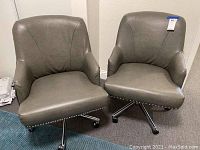 Two gray rolling desk chairs side by side, showing front and angled sides, with curved armrests, padded seat and back, and decorative nailhead trim at base of seat.