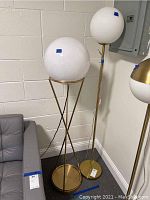 Two modern floor lamps with white spherical globe shades, brass-colored metal stands and bases, positioned next to a gray sofa against a white brick wall.