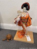 Full view of the Geisha figurine and wooden frog on carpeted floor against white wall.