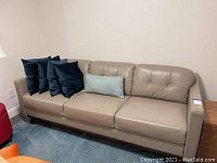 Full view of taupe leather sofa with five throw pillows (four dark blue, one light blue). Sofa has tufted back cushions and wooden legs.