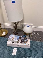 Photo showing modern branch-style lamp with white fabric shade, modern spherical lamp with brass and marble base, and boxes of LED light bulbs on carpet.